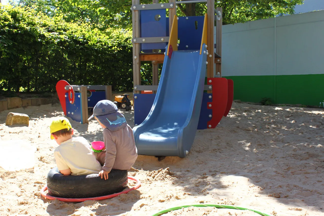 Kinder spielen im Sandkasten des Außengeländes, im Hintergrund eine blaue Rutsche und ein Klettergerüst.