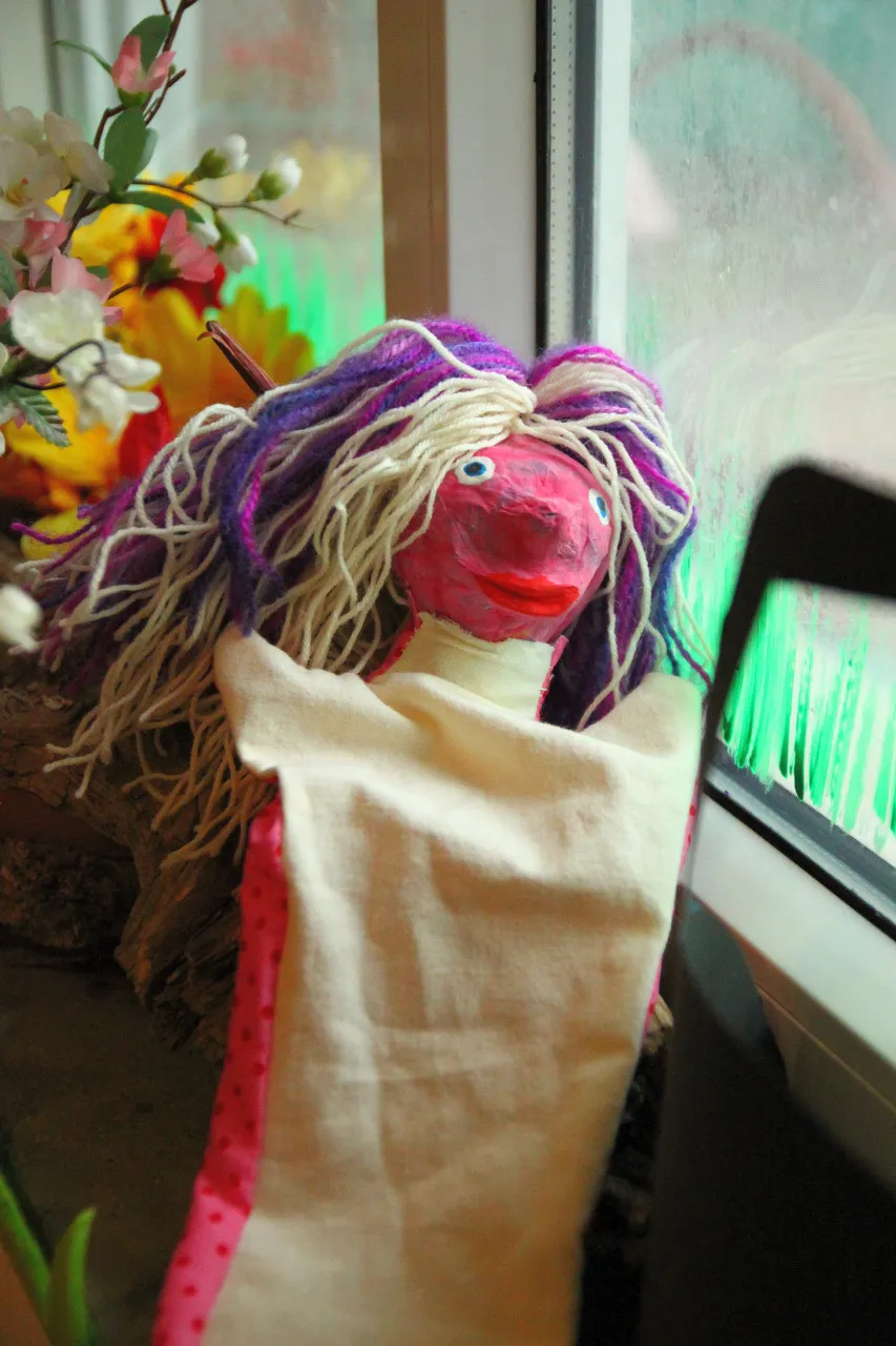 Handpuppe mit gelbem Wollhaar vor einer Fensterbank mit Blumen.