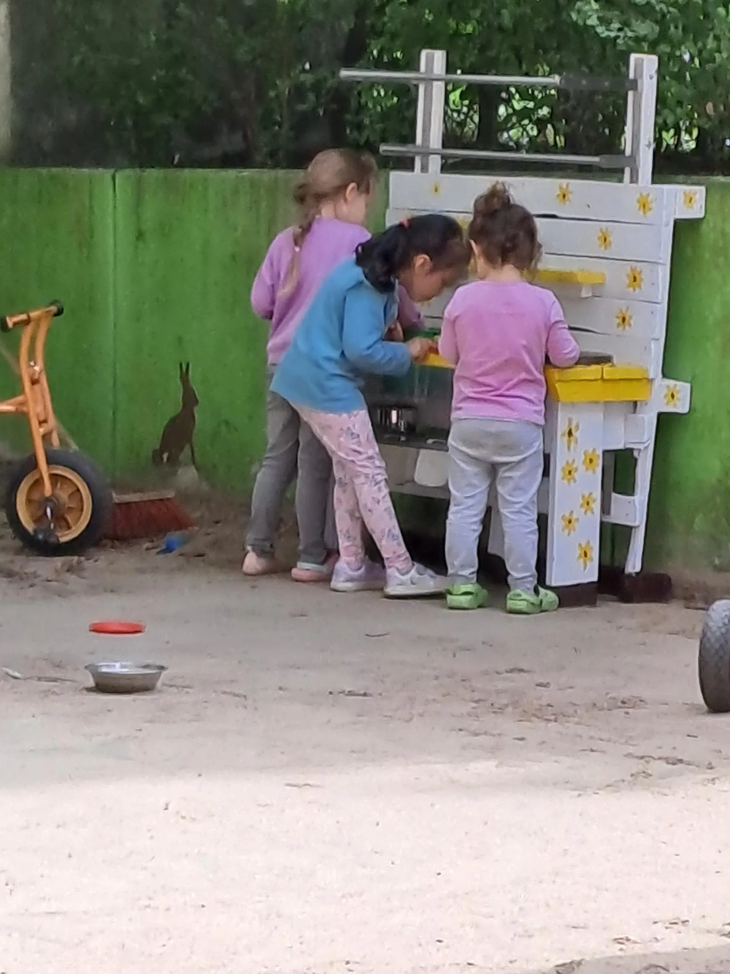 Drei Kinder stehen gemeinsam an der Matschküche, daneben ein Dreirad im Sand.