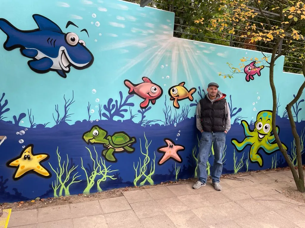 Außenwand des Kinderladens, besprüht mit einer bunten Unterwasserwelt aus Hai, Fischen, Schildkröte, Seesternen und einem Tintenfisch. Davor steht ein Mann mit Mütze und Weste.