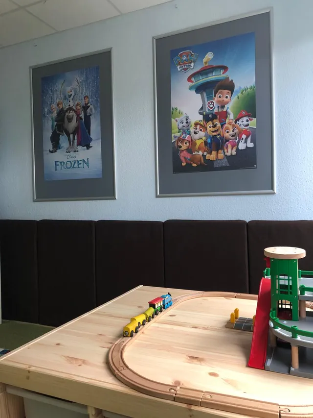 Heller Bauraum mit einem großen Holztisch, auf dem eine Holzeisenbahn mit Zug und Parkhaus aufgebaut ist. An der hellblauen Wand hängen zwei gerahmte Kinderposter.